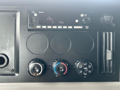 Dashboard panel of a Freightliner Cascadia showing climate controls, radio/FM2 tuner, and power outlet in a sleeper truck interior.