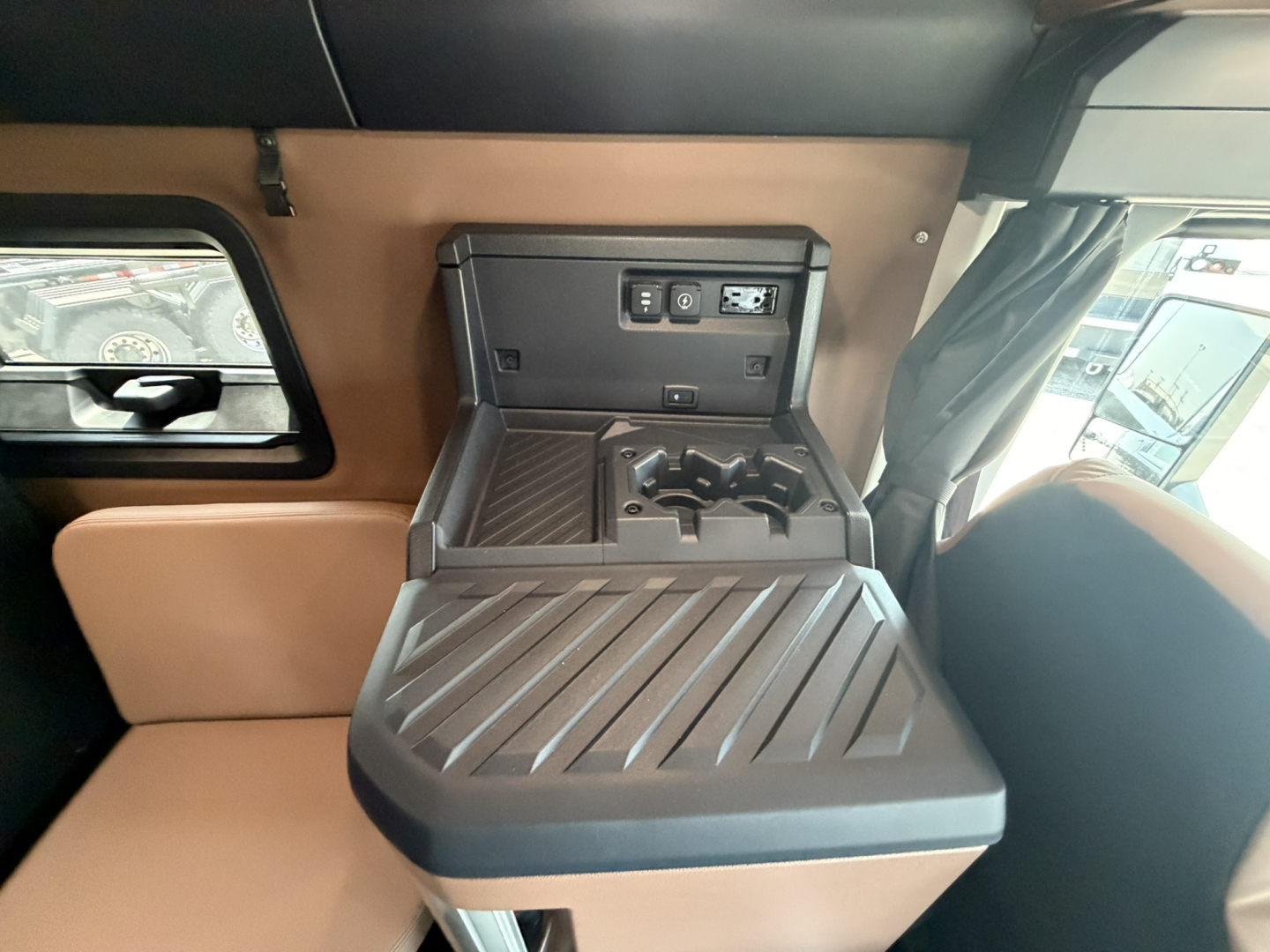 Cab interior showing center console with cupholders and control panel, tan seating and window with side mirror in view.