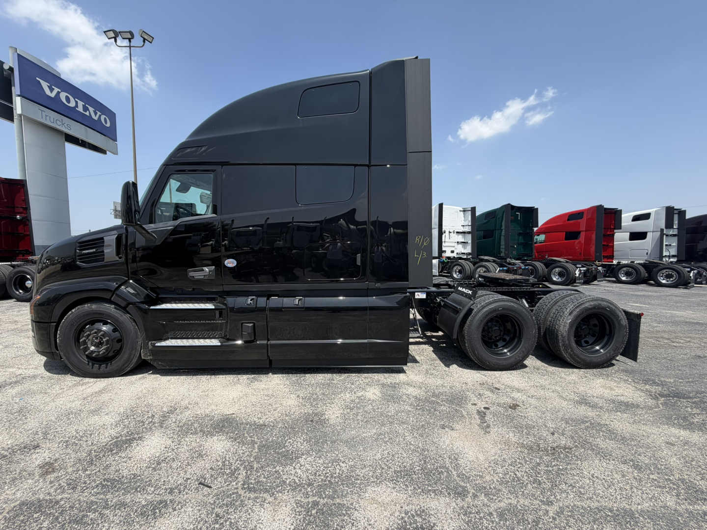 Mack PR64T76H conventional sleeper truck in black, side view showing cab and rear axles, with Volvo dealership sign in background.