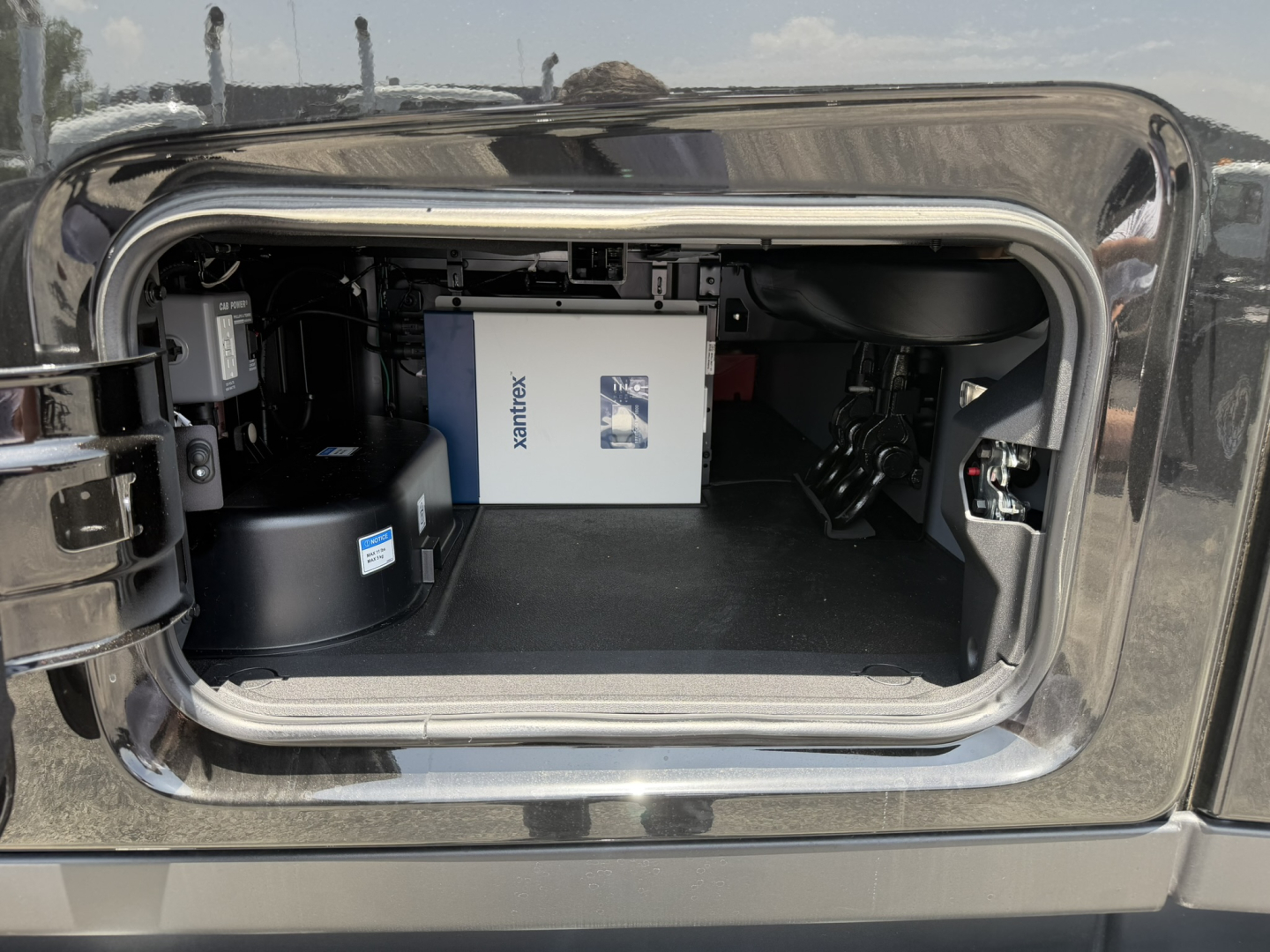 Truck cab rear hatch open showing storage area with a white box labeled MACK? and a blue label, auxiliary cabinet, and black components inside the sleeper compartment.