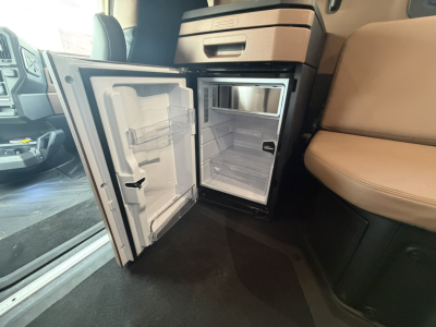 Mack truck cab interior showing open compact refrigerator/freezer mounted under dash in sleeper area and passenger seat beside; interior is light tan with storage compartments.