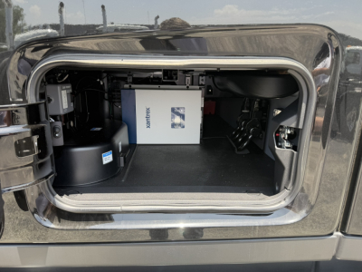 Truck cab rear hatch open showing storage area with a white box labeled MACK? and a blue label, auxiliary cabinet, and black components inside the sleeper compartment.