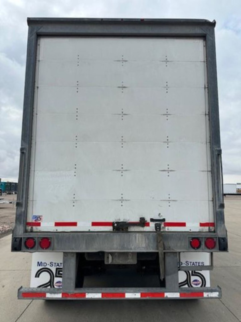 Rear of a white Wabash trailer with doors closed; rear lights and bumper visible; reflective tape along bottom; small metal latch and hinge hardware; license/registration sticker areas.