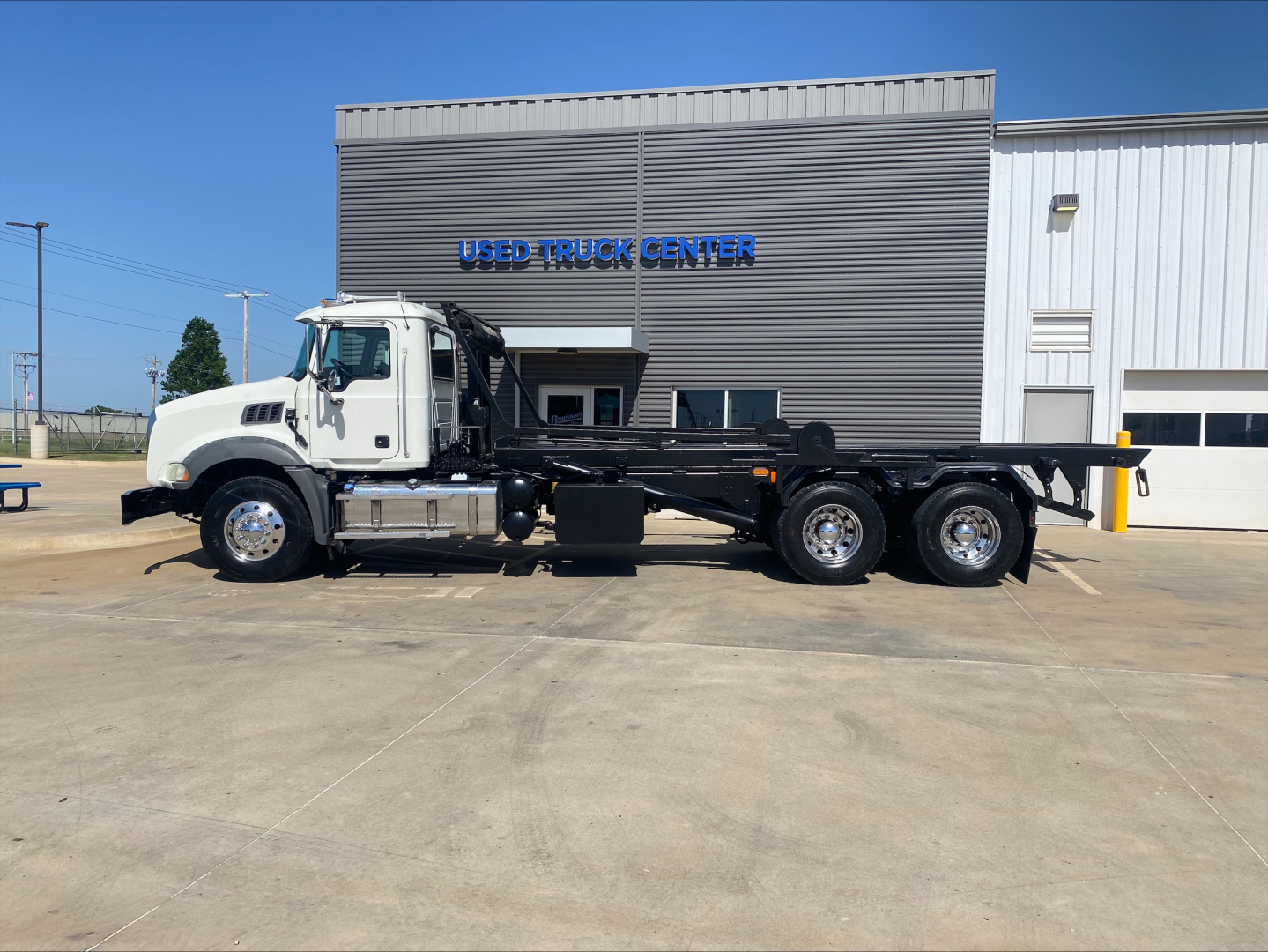Vehicle side view of a 2016 Mack GU533 roll-off truck showing chassis and rear axles; building with blue 'USED TRUCK CENTER' sign in background.
