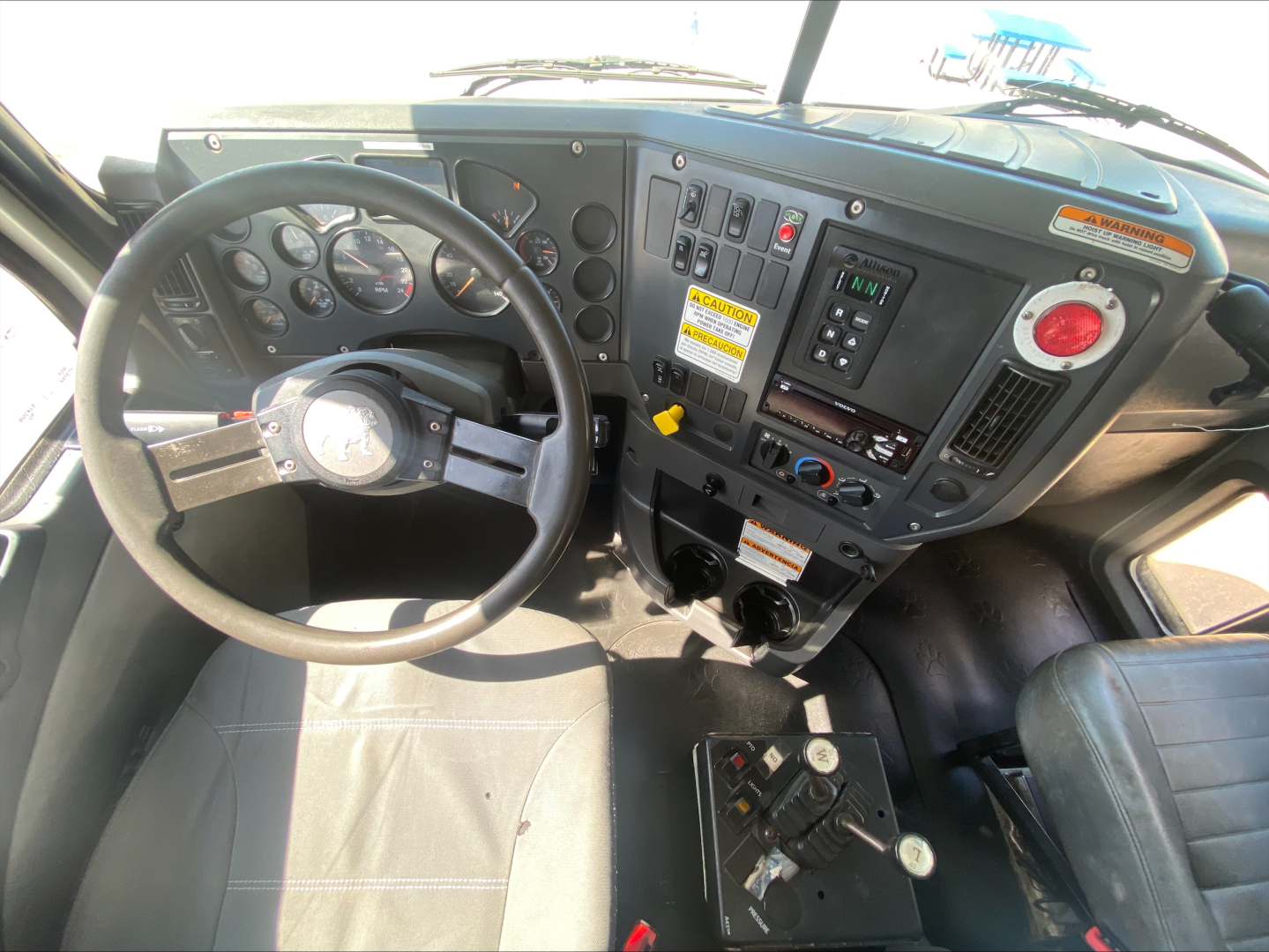 Cab interior of a 2016 Mack GU533 roll-off truck showing steering wheel, dashboard with gauges, controls, and gear selector.