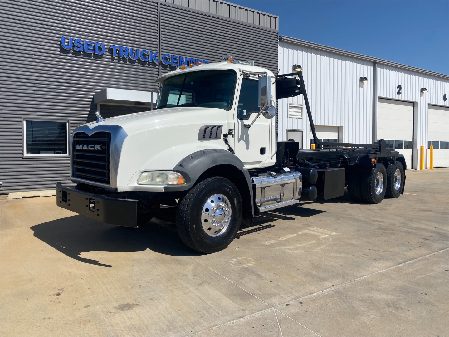 Mack GU533 heavy-duty truck in white, side view showing roll-off chassis and hydraulic boom, parked in front of Used Truck Center building.