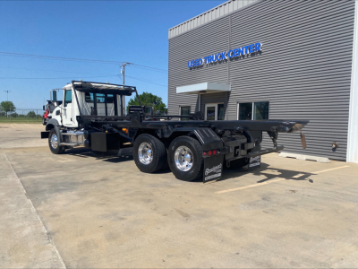 Flatbed/rolloff truck with a Mack GU533 cab in front, chassis visible, at a truck center. Visible text includes 'TRADE TRUCK CENTER' on the building.