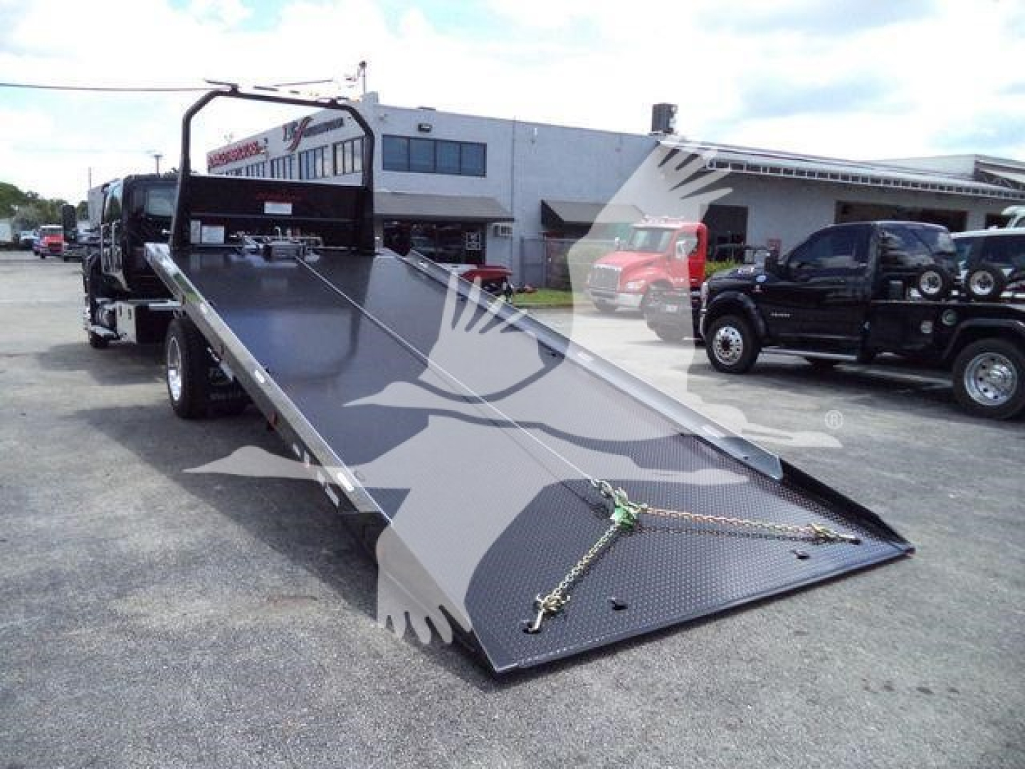 Truck bed shows a ramped rollback trailer with a black steel surface and diamond plate ramp. It has a winch line and hooks at the front, with a cab-over truck in background.