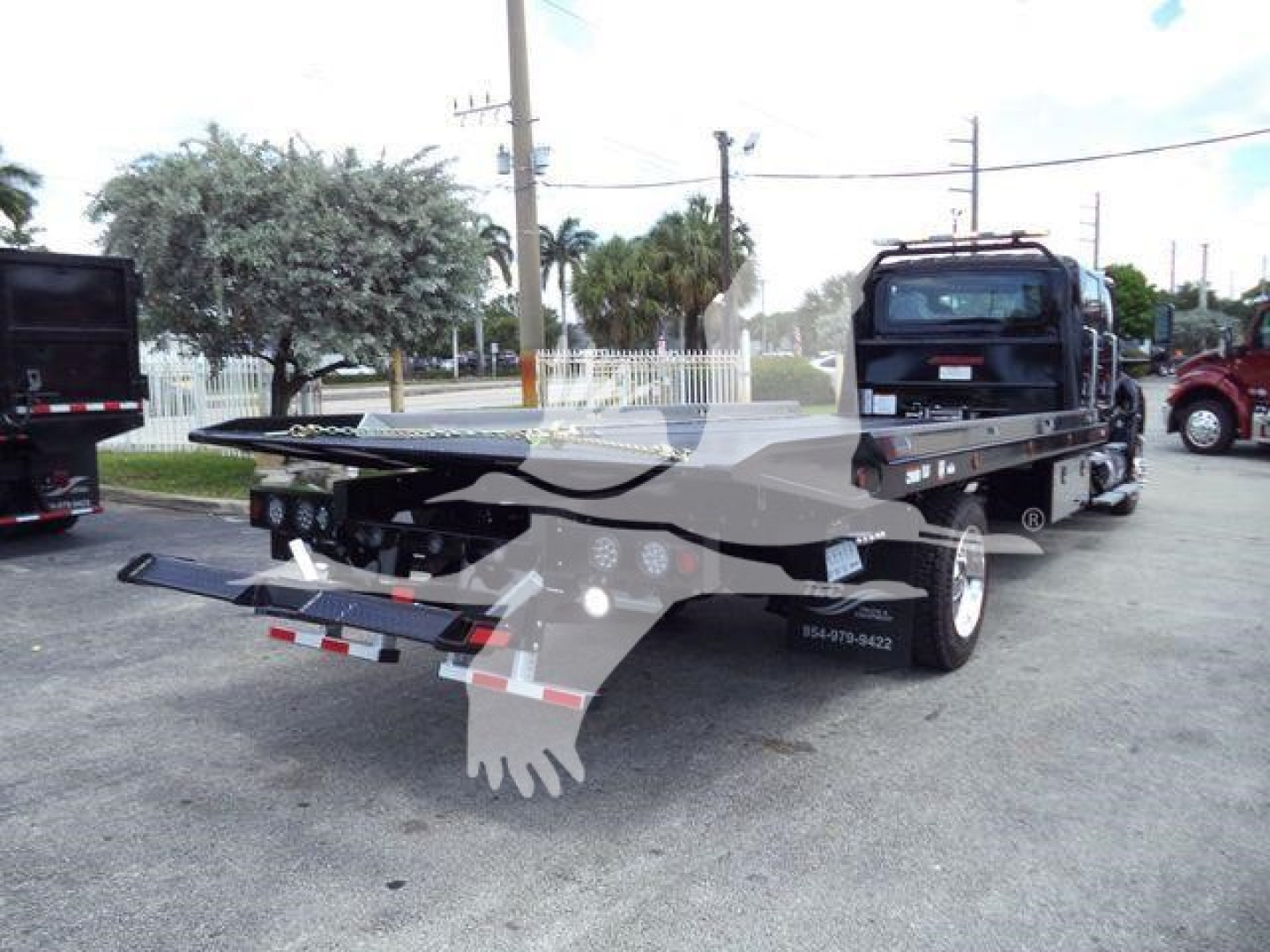 Tractor tow-roller truck with flatbed in a parking lot. Visible text on the side panel shows 'BMVCREW22SRR6T' and contact digits. The background includes other trucks and a white fence.