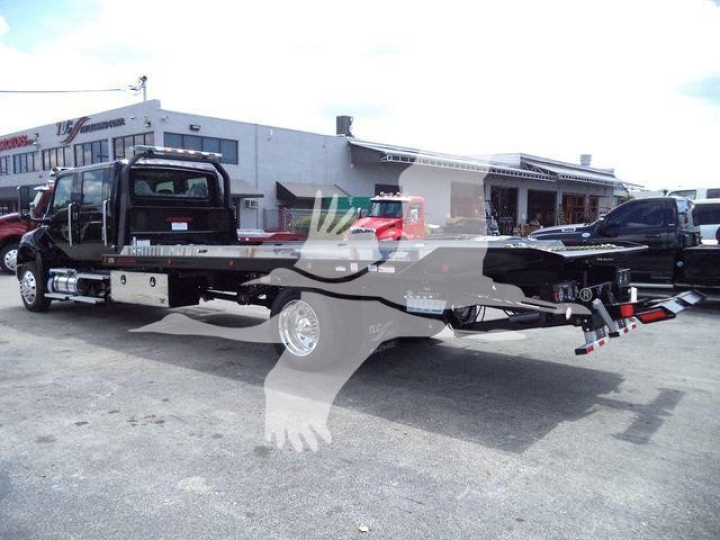 Flatbed tow truck in a dealership lot showing a long tow bed with wheel lift and dual rear axles, International MV chassis, side rails, and rear hitch components.
