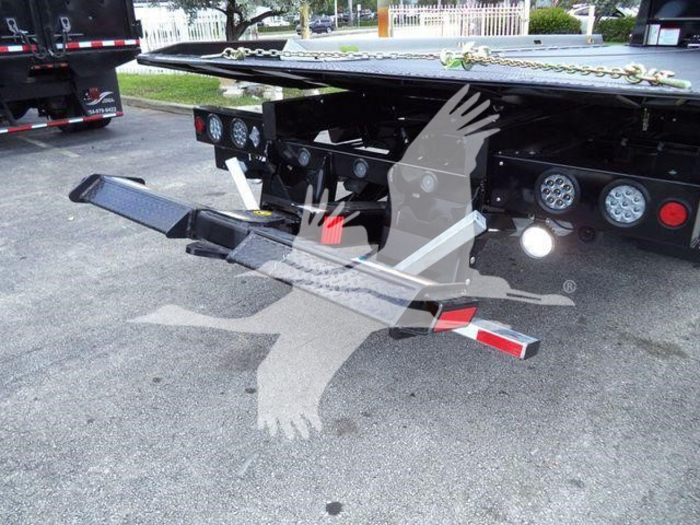 Flatbed tow truck rear with black metal frame, multiple round taillights, and a dual-sided metal ramp; chain visible on top; no legible brand text; in-service equipment area.
