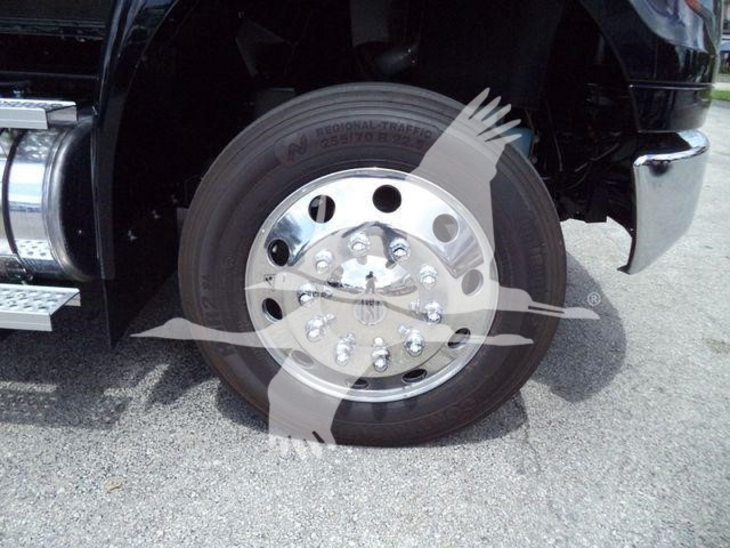Truck front left wheel and fender showing aluminum hub and tire, chrome step bumper, and black drivetrain area.
