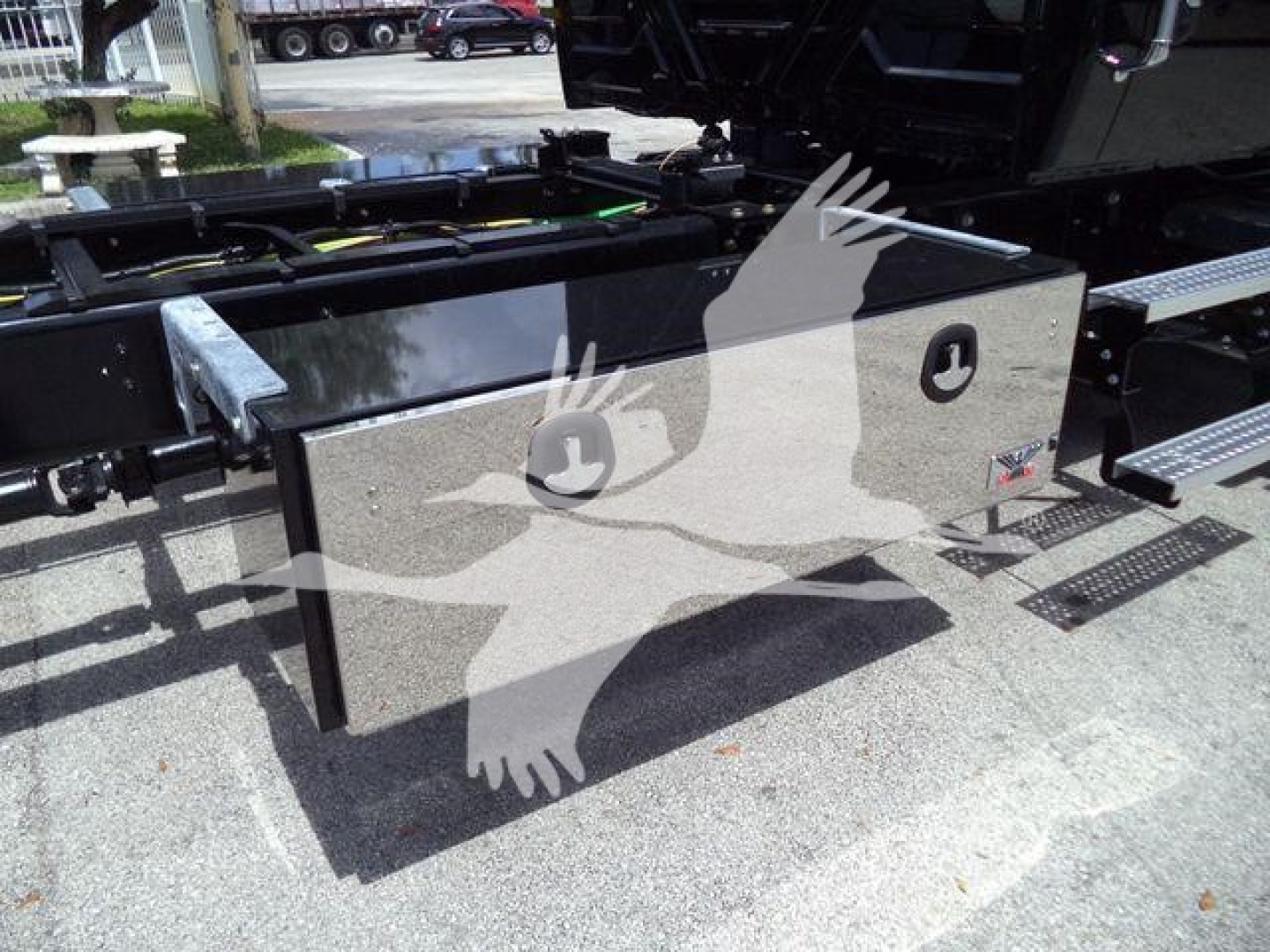 Truck equipment box mounted on chassis showing metal toolbox with two circular latches and a small red/white sticker on the side.