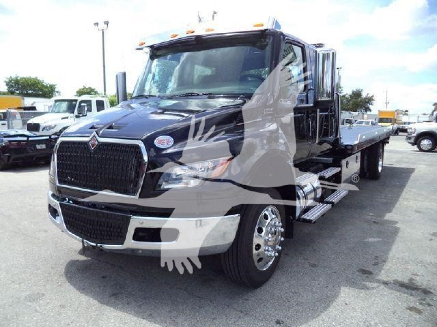 Truck cab of a 2026 International MV rollback truck showing front grille, headlights, and side steps with a flatbed in a dealership lot.