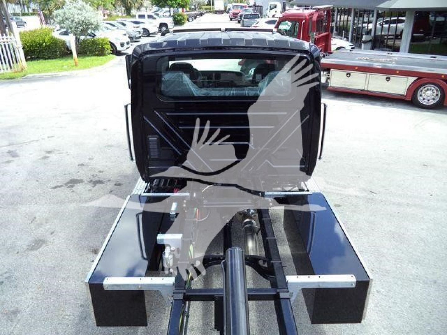 Truck chassis with hydraulic rollback hitch and damaged cab in background; matte black metal components and control leverage visible.