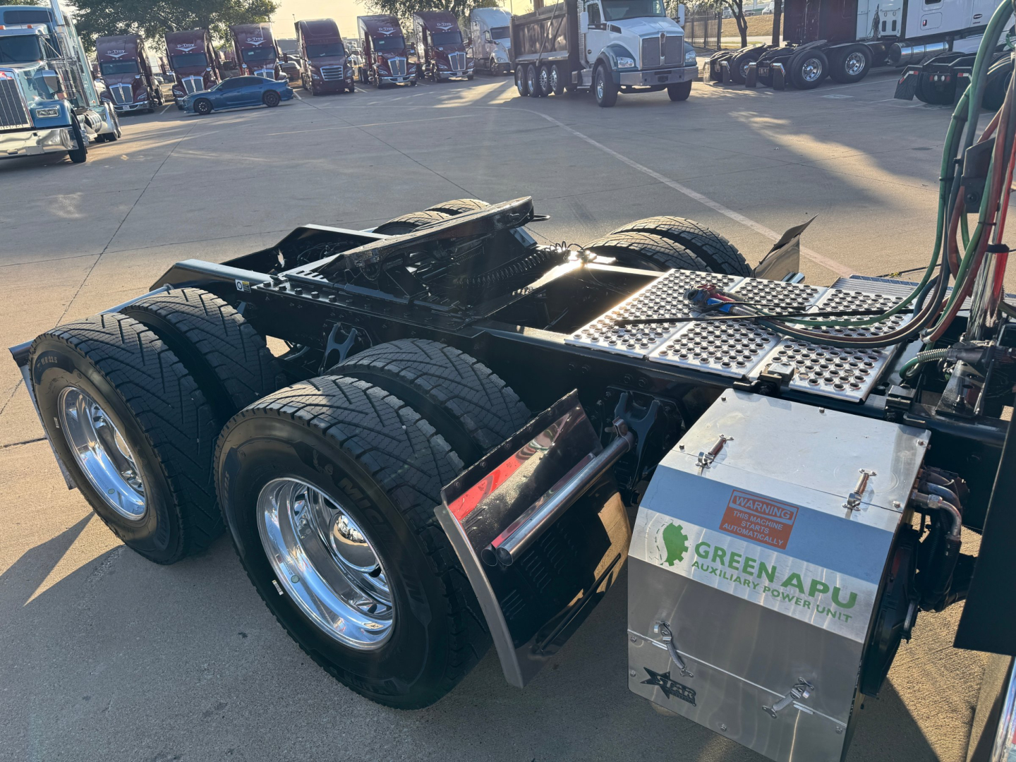 Kenworth W900L chassis showing rear dual-axle configuration and exposed aluminum toolbox with Green APU sticker; multiple trucks visible in background.