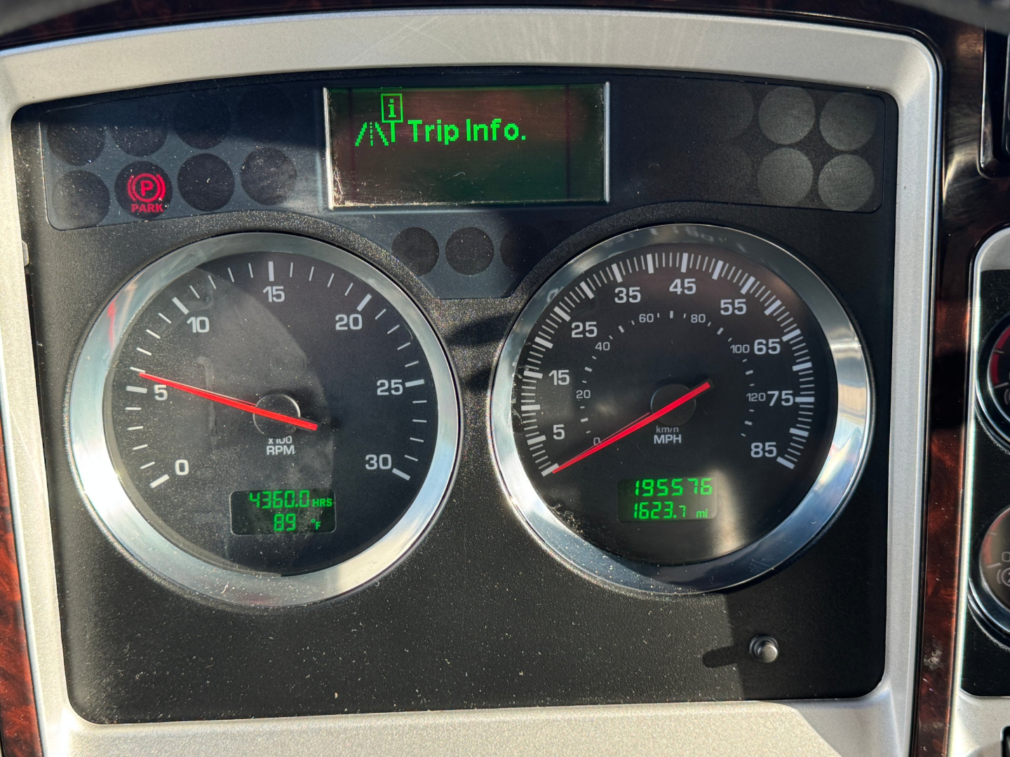 Kenworth truck instrument cluster showing circular gauges, left RPM gauge, right speedometer, digital trip info screen above, green/readouts on lower panels.