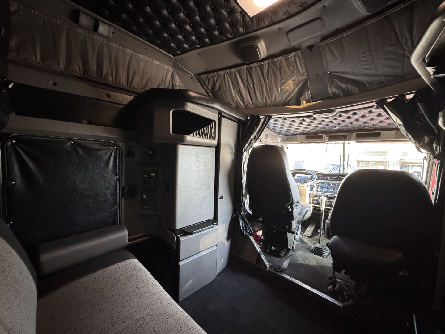 Cab interior of a Kenworth W900L showing driver and passenger seats, dashboard, and sleeper area with overhead cabinets.