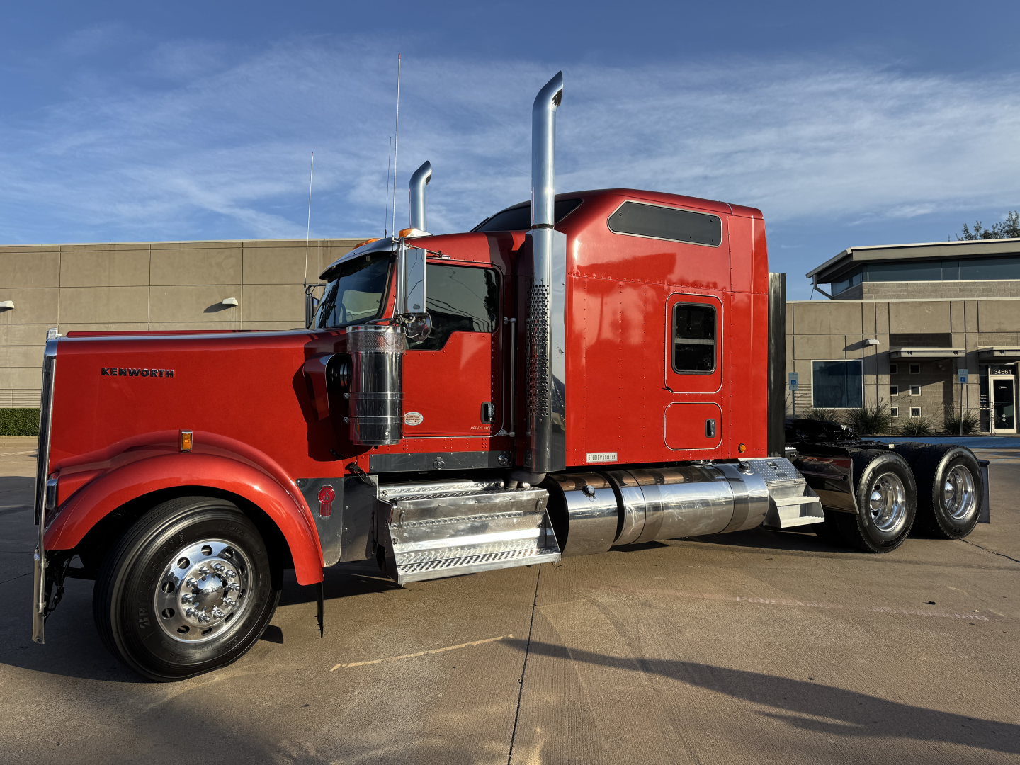 Kenworth W900L truck in red, side view showing chrome exhaust stacks, fuel tanks, and heading cab with windows. Visible text includes KENWORTH on the side of the hood.