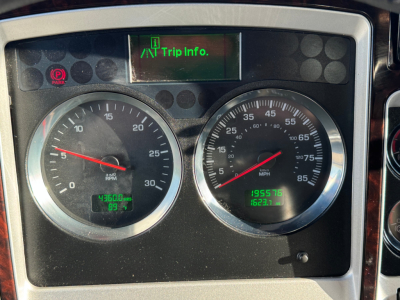 Kenworth truck instrument cluster showing circular gauges, left RPM gauge, right speedometer, digital trip info screen above, green/readouts on lower panels.