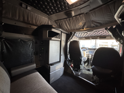 Cab interior of a Kenworth W900L showing driver and passenger seats, dashboard, and sleeper area with overhead cabinets.