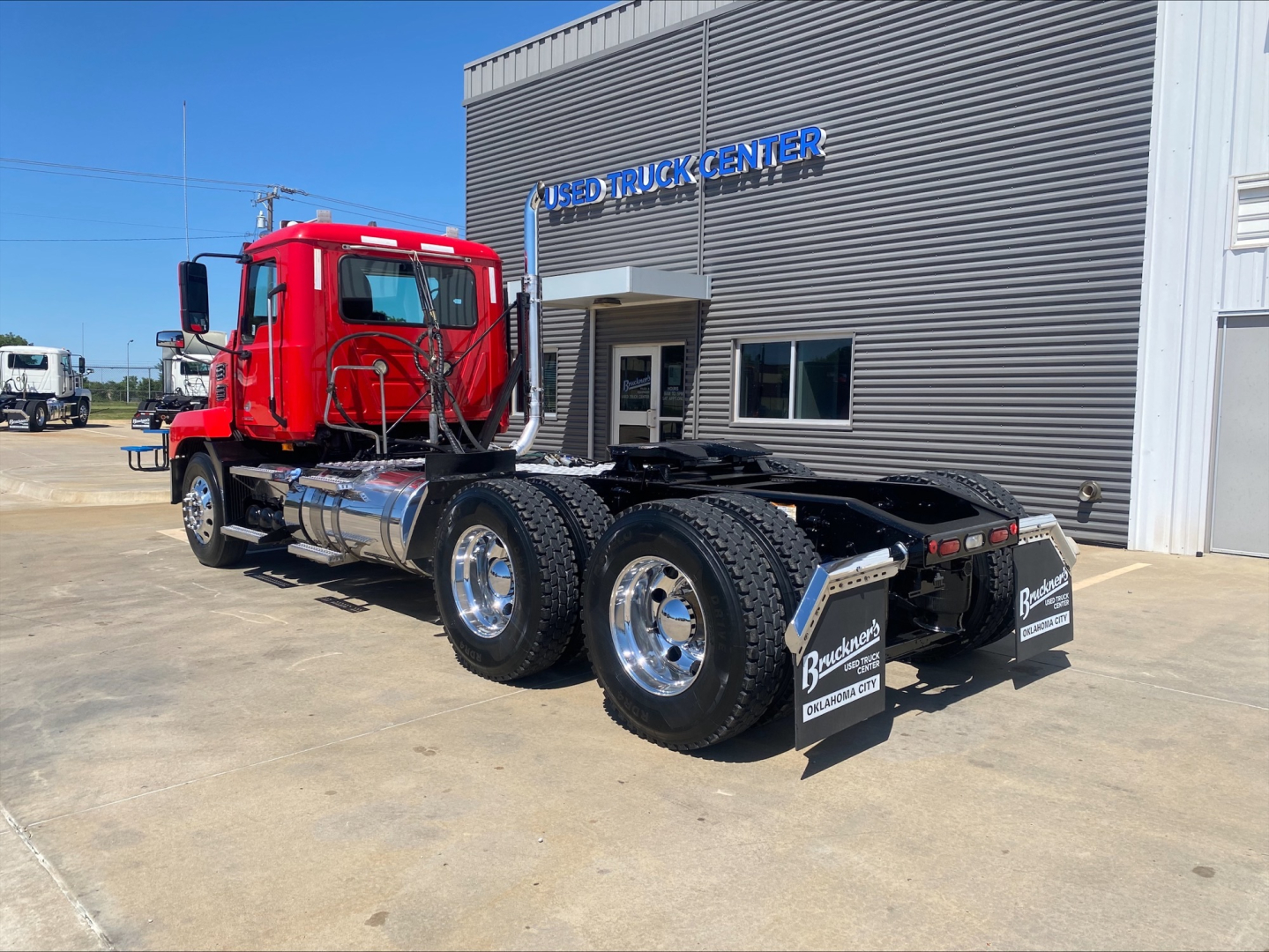 Red 2021 Mack Anthem daycab truck with chrome fuel tanks and dual rear axles parked outside a building labeled Used Truck Center.