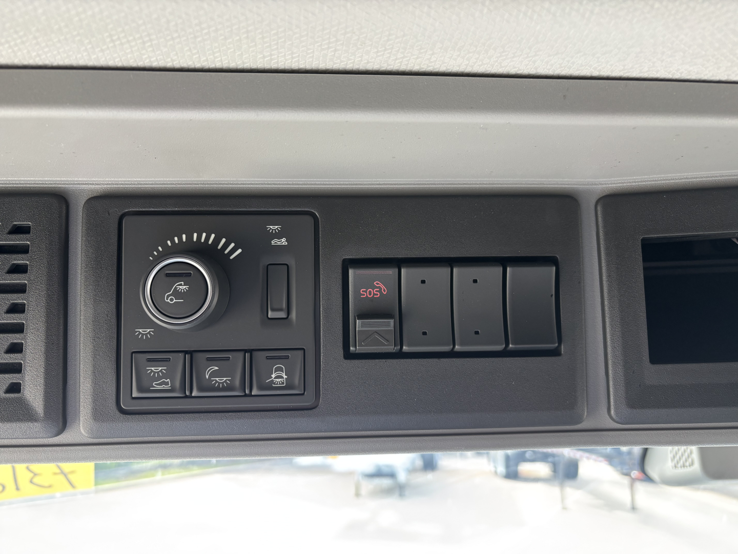 Dashboard panel with control dial and several switches in a Volvo VNL, including circular knob with icons, rectangular buttons, and a 5-switch block labeled SOS.
