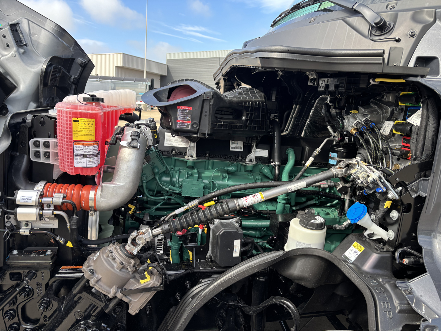 Volvo VNL64T860 engine bay view showing green engine block, red coolant reservoir, various hoses and wiring harnesses, and plastic fluid reservoirs.