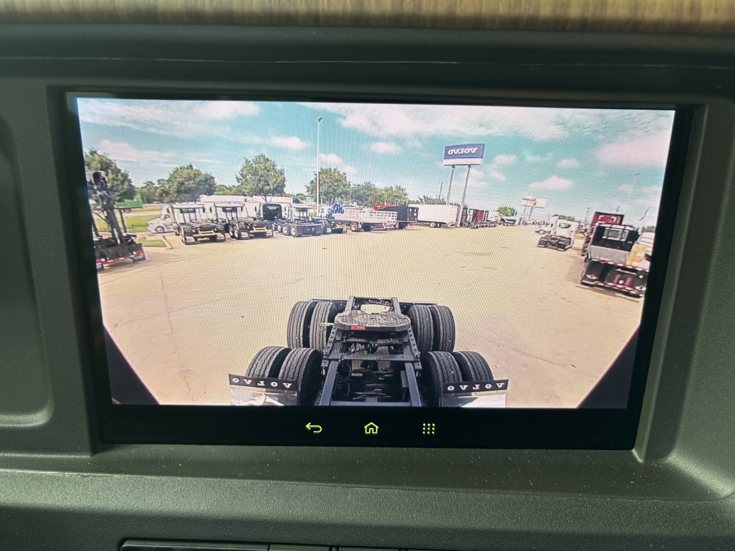 Rear cab view of a Volvo VNL truck chassis in a lot, with a drive axle and fifth wheel visible; building and other trucks in background.