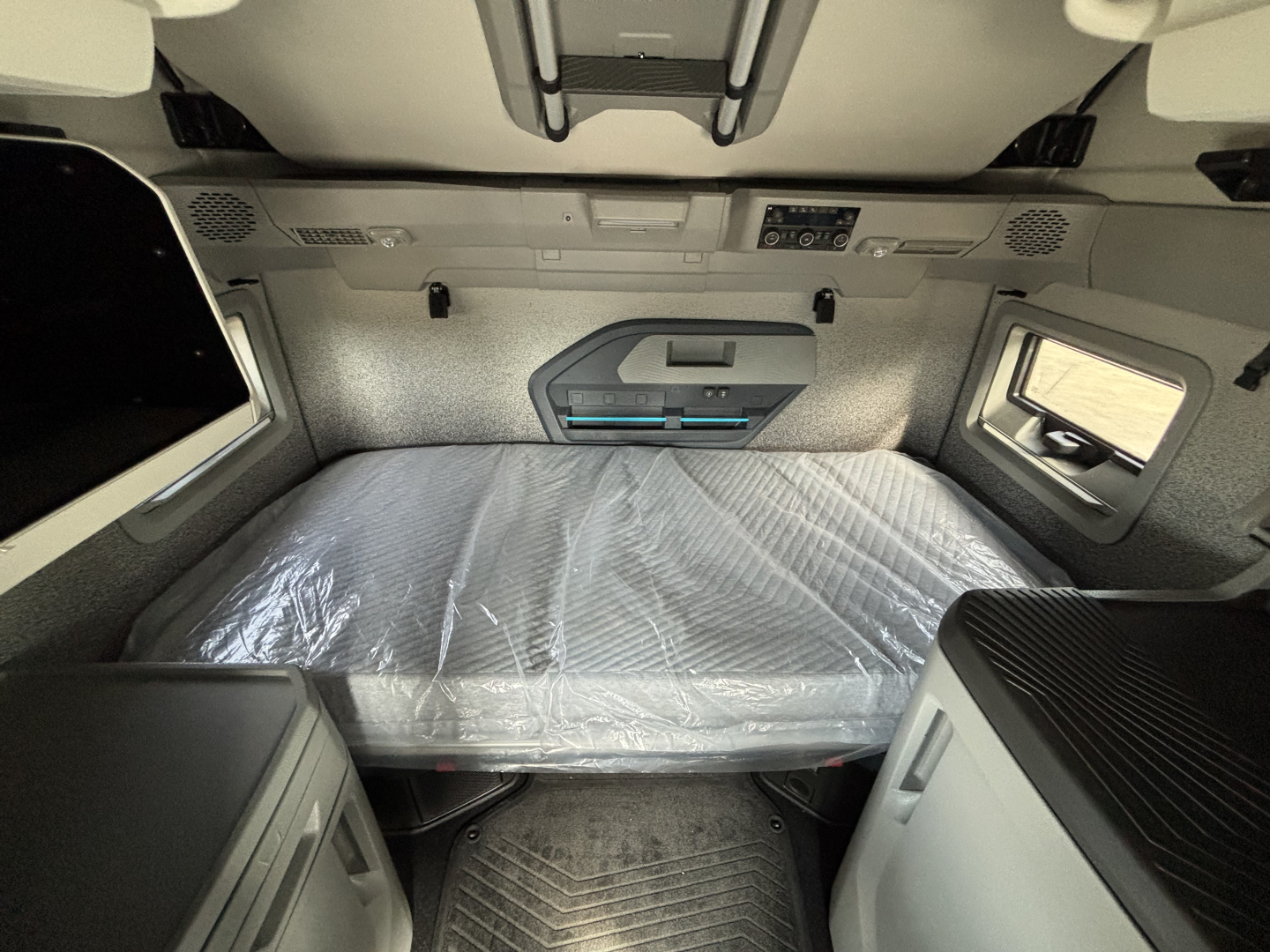Volvo VNL64T860 sleeper cab interior showing bed with plastic-wrapped mattress, overhead storage panels, side window, and control panel on rear wall.