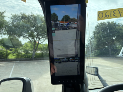 Interior rear-view mirror/monitor in a Volvo VNL truck showing a digital display with exterior yard scene reflected, windshield with trees and vehicles.