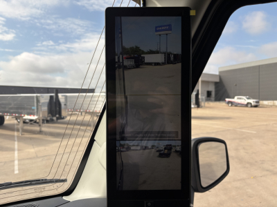 Truck cabin interior with a tall touchscreen tablet mounted near the windshield; exterior lot with trucks and a building visible in the background.