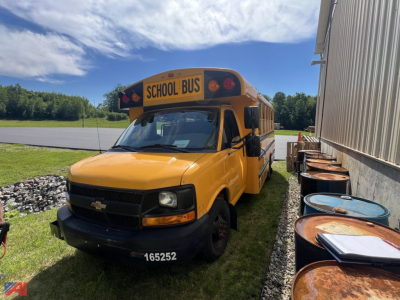School bus in front of a building, yellow Chevrolet Express school bus with 'SCHOOL BUS' sign on the roof, front license/ID 165252, daytime outdoor setting.