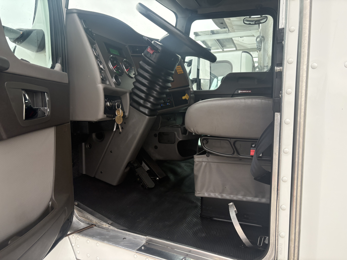 Cab interior of a Kenworth T270 truck showing steering column, dashboard, pedals, and driver seat; left door open, floor mat visible.