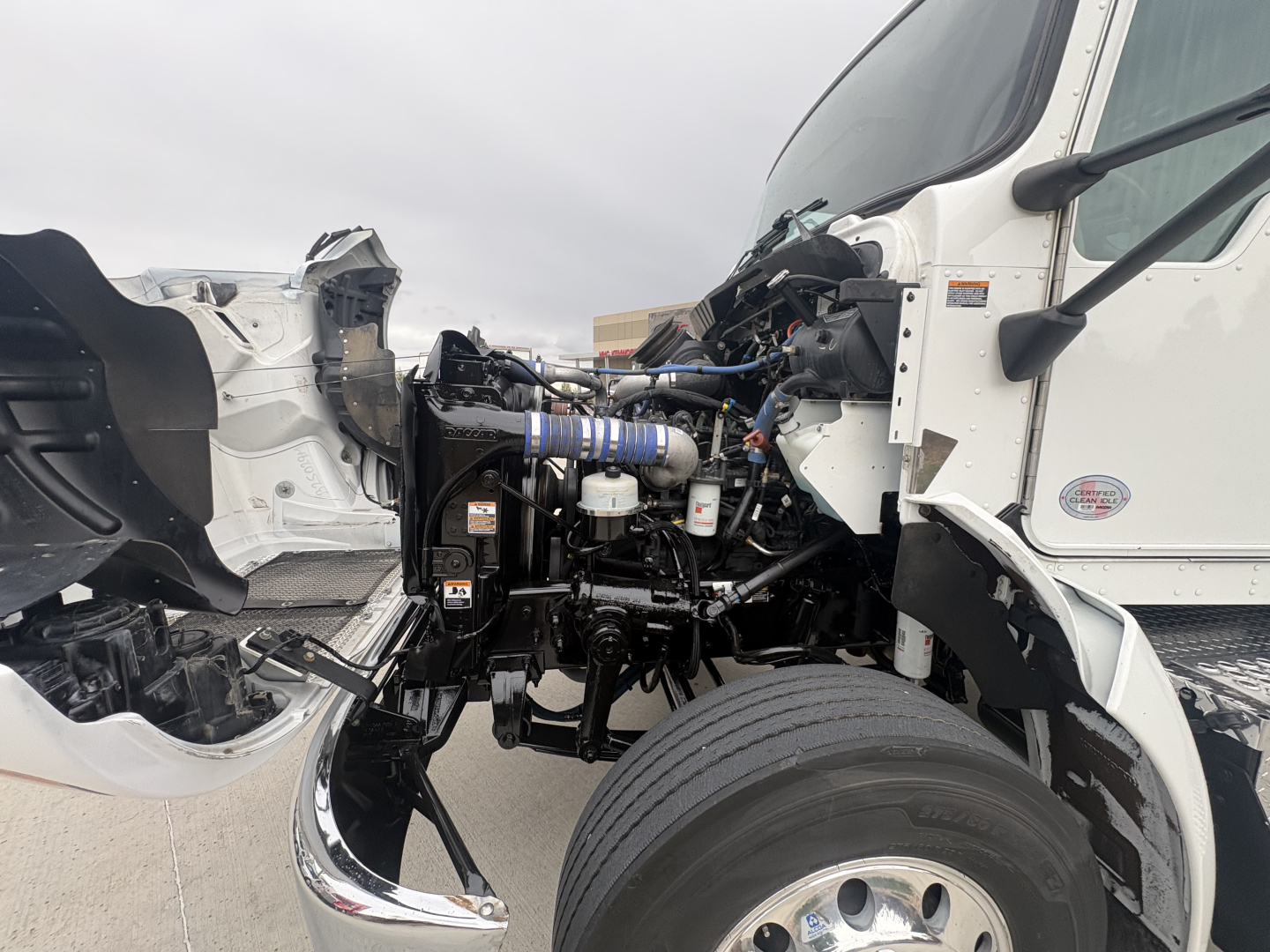Kenworth truck engine bay view with exposed frame and components showing engine, hoses, and front wheel; cab removed near engine bay.