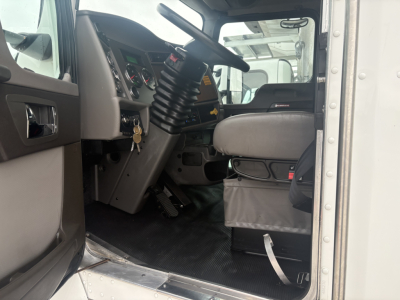 Cab interior of a Kenworth T270 truck showing steering column, dashboard, pedals, and driver seat; left door open, floor mat visible.