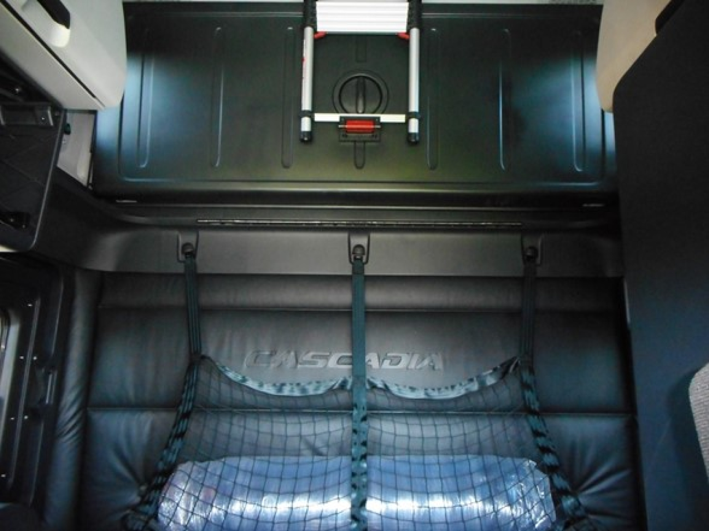 Truck cab interior showing Freightliner Cascadia sleeper area with seats and cargo net, view toward rear of cabin.