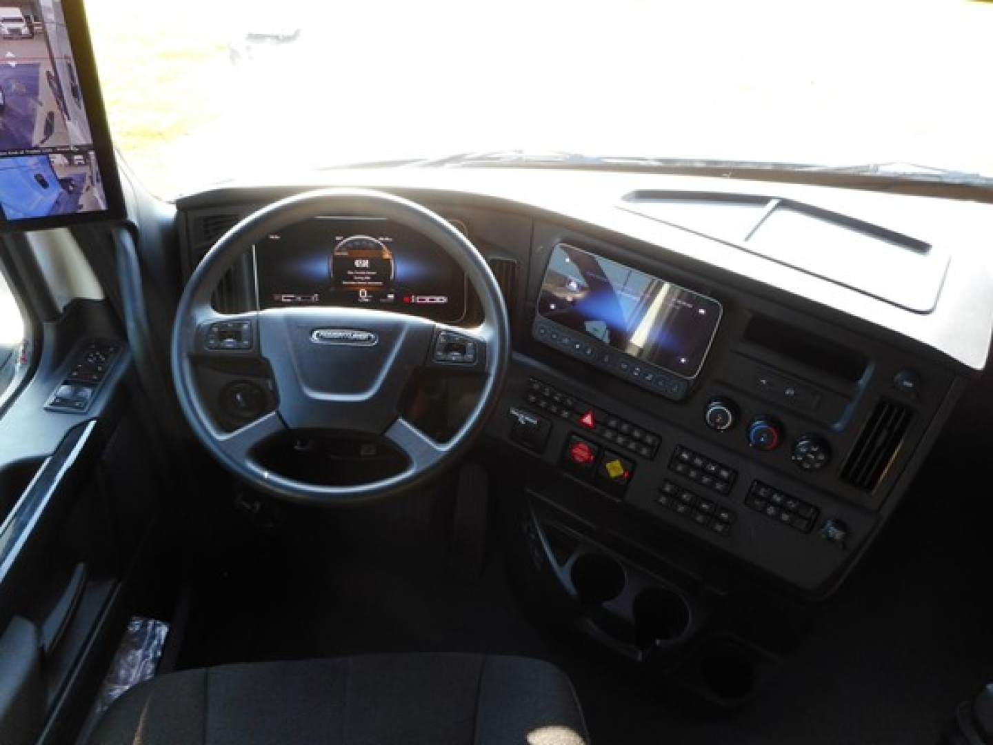 Inside view of a 2026 Freightliner Cascadia 126 cab: steering wheel, dashboard with digital gauge, large infotainment screen, multiple control panels, cup holders.