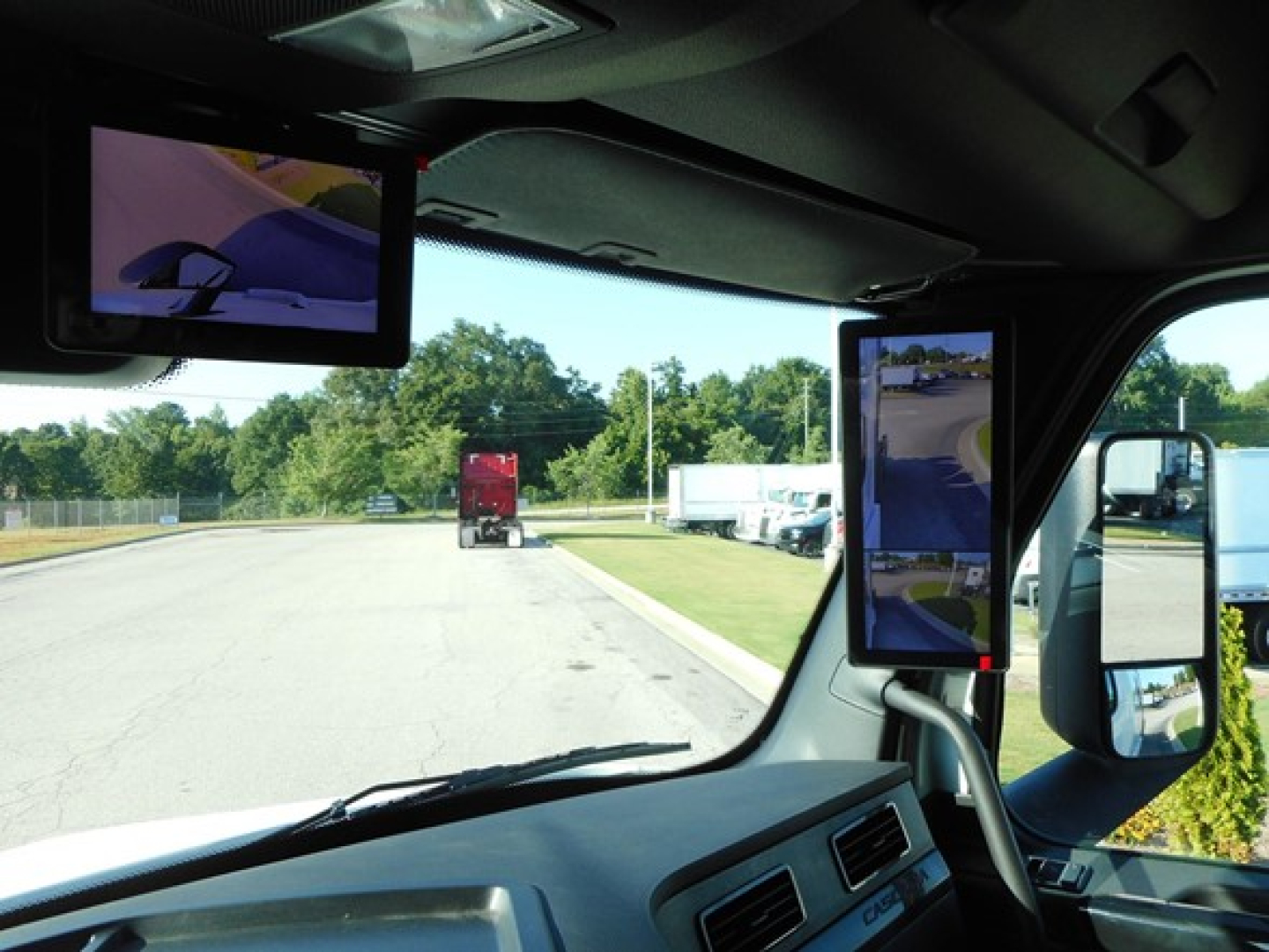 Interior cab view of a 2026 Freightliner Cascadia 126 with multiple screens mounted inside, side mirror visible, road and truck in background.