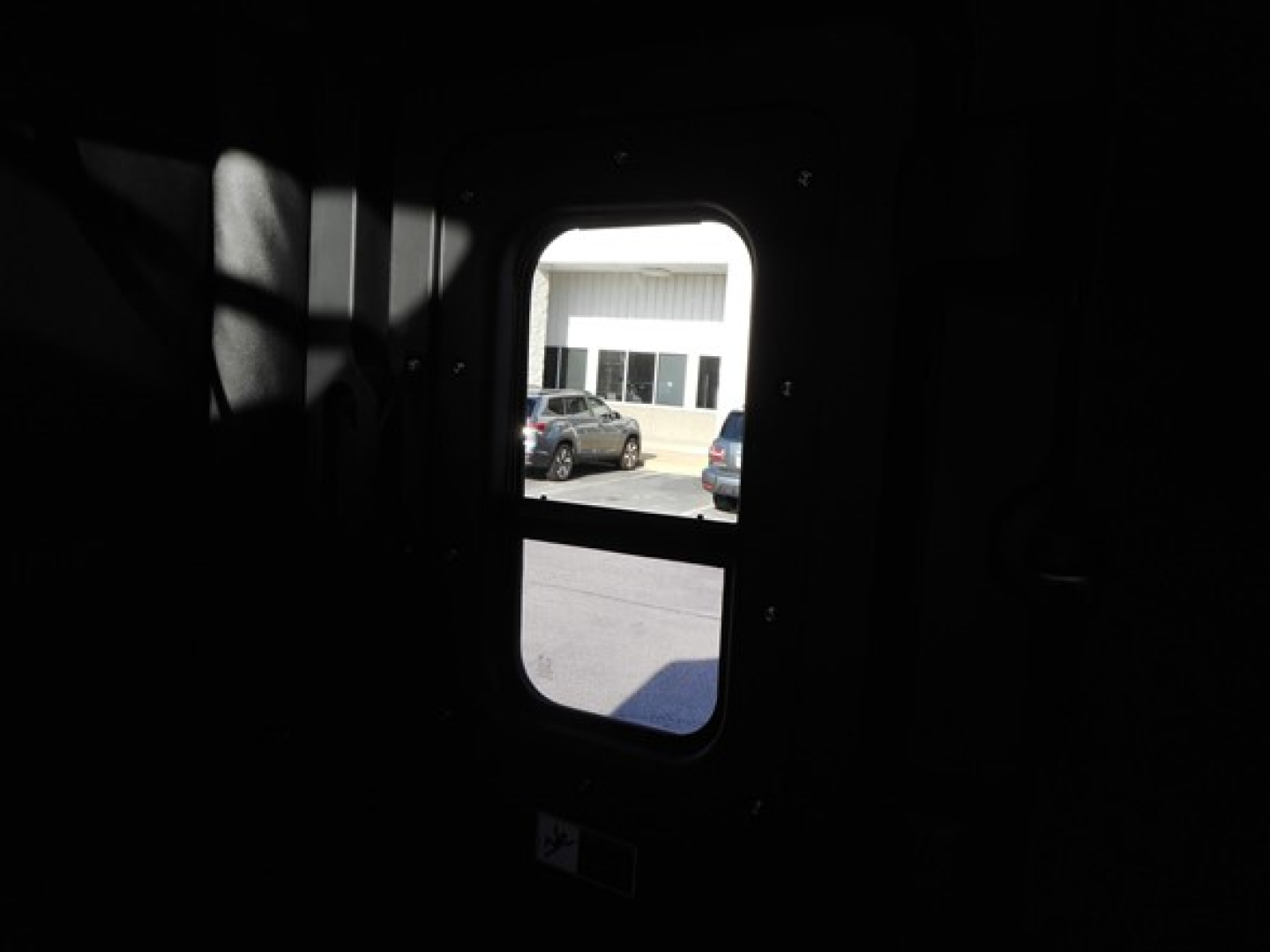 Truck cab window view showing exterior scene through small rectangular door window, view into daylight outside with parked vehicles.