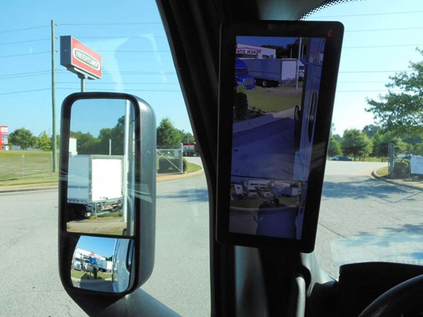 Freightliner Cascadia cab interior view showing side mirror and vertical monitor with reflected outdoor lot.
