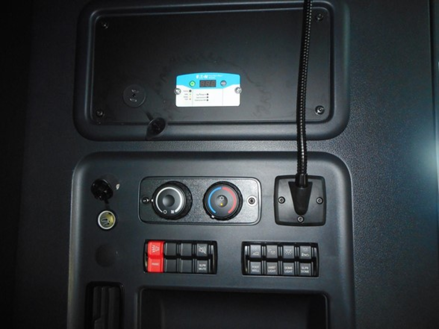 Dashboard panel of a Freightliner Cascadia showing climate controls, switches, and a circular dial with a blue sticker plate above. Includes a microphone cord and various control buttons.