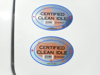 Two circular Certified Clean Idle stickers on a white truck door, blue border with orange center, text reads CERTIFIED CLEAN IDLE. Detroit and state codes visible.