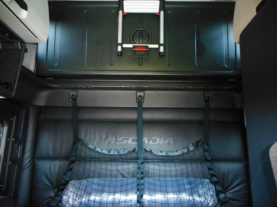 Truck cab interior showing Freightliner Cascadia sleeper area with seats and cargo net, view toward rear of cabin.