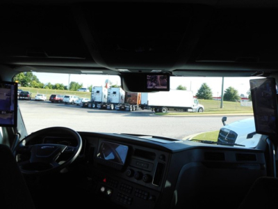 Cab interior of a 2026 Freightliner Cascadia 126 tractor showing dashboard and steering wheel with large side mirrors and windshield view of a truck yard.