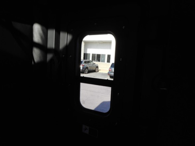 Truck cab window view showing exterior scene through small rectangular door window, view into daylight outside with parked vehicles.