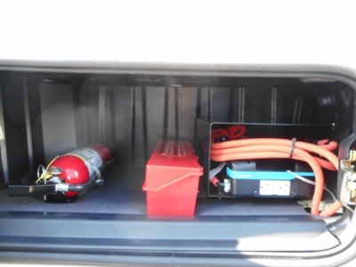 Freightliner Cascadia 126 engine bay area showing battery, orange cables, red battery box, and a red fire extinguisher canister.