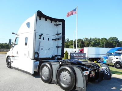 Truck tractor in a lot with a white Freightliner Cascadia, open cab rear view showing sleeper area and rear wheels. signage reads PEACH STATE TRUCK CENTERS and American flag on pole; other trucks visible in background.