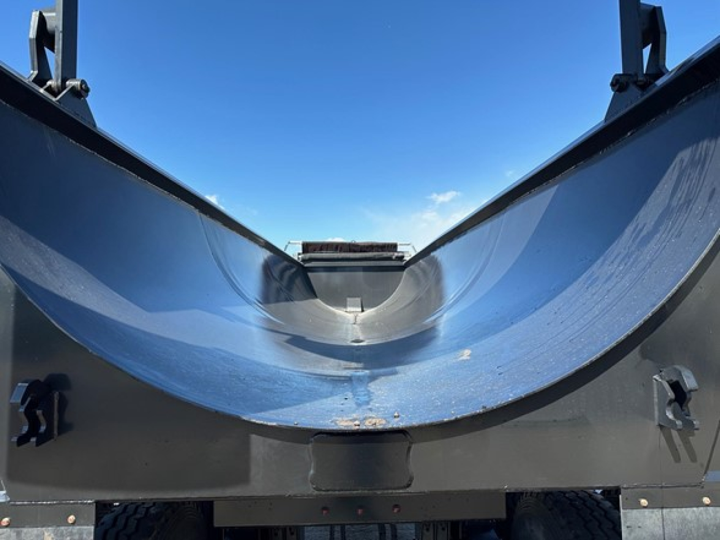 New dump trailer cargo bed showing curved rock tub with steel sidewalls and underside, reflective blue interior surface, rear hitch area visible.