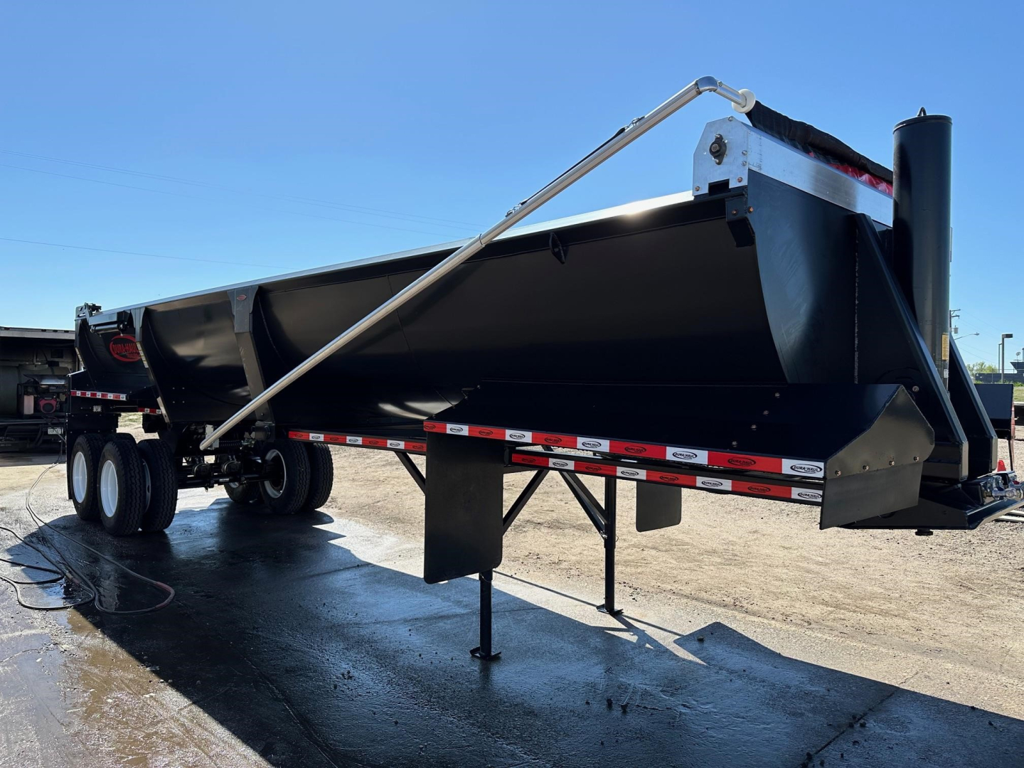 End dump trailer with frameless steel rock body, high lift gate, and tri-axle wheels, showing hydraulic lift mechanism and rear reflective strips.